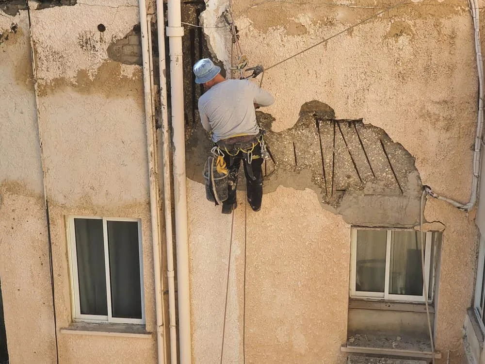 עובד עבודות גובה בסנפלינג מבצע שיקום קיר חיצוני בבניין ישן, עם טיפול באזורי טיח ובטון מתפוררים, ברזל חשוף וחזית פגועה לצד חלונות וצנרת חיצונית. עובד עבודות גובה בסנפלינג מבצע שיקום קיר חיצוני בבניין ישן, עם טיפול באזורי טיח ובטון מתפוררים, ברזל חשוף וחזית פגועה לצד חלונות וצנרת חיצונית.